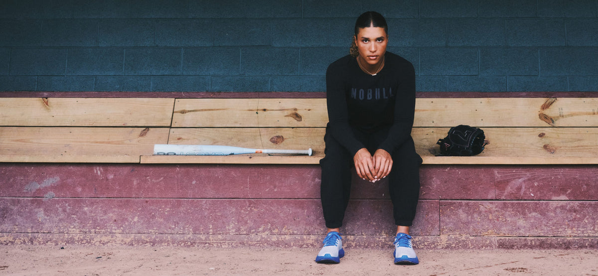 Maya Beady sits on wooden stadium bleachers wearing a black long-sleeve top with 'NOBULL' printed across the chest, black pants, and blue sneakers. A baseball bat rests on the bench beside them, with a dark brick wall behind.