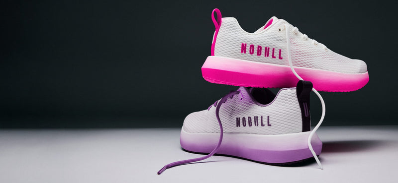 A pair of white athletic sneakers with vibrant pink and purple gradient accents, stacked with one shoe floating above the other against a dark gray background. The brand name 'NOBULL' is printed on the side of the shoes.