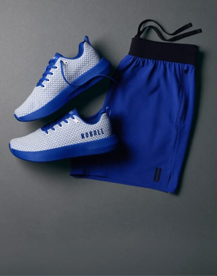 A pair of white and blue athletic sneakers next to a pair of royal blue shorts with a black waistband, arranged on a dark gray background. The brand name 'NOBULL' is visible on the side of the shoe.