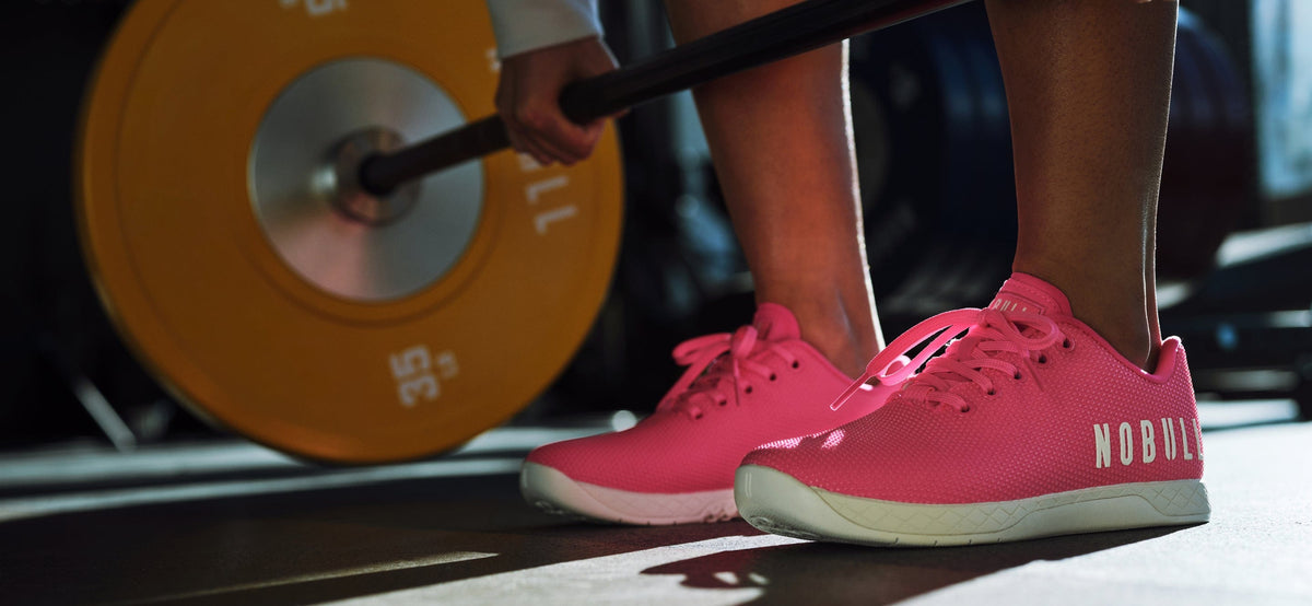 Person lifting a barbell with pink Outwork Shoes on a gym floor