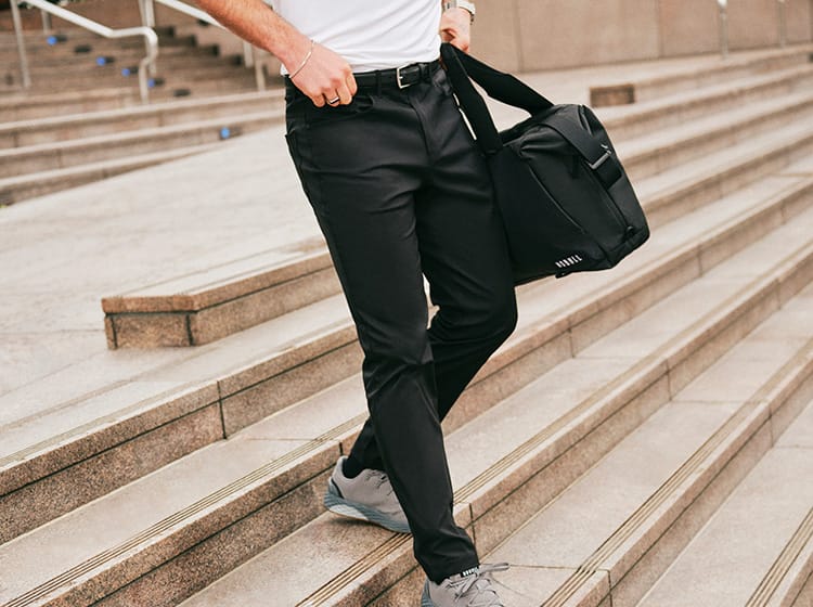 A person walking down outdoor stone steps while carrying a black duffel bag over one shoulder. The person is wearing black trousers, a white top, and gray sneakers.
