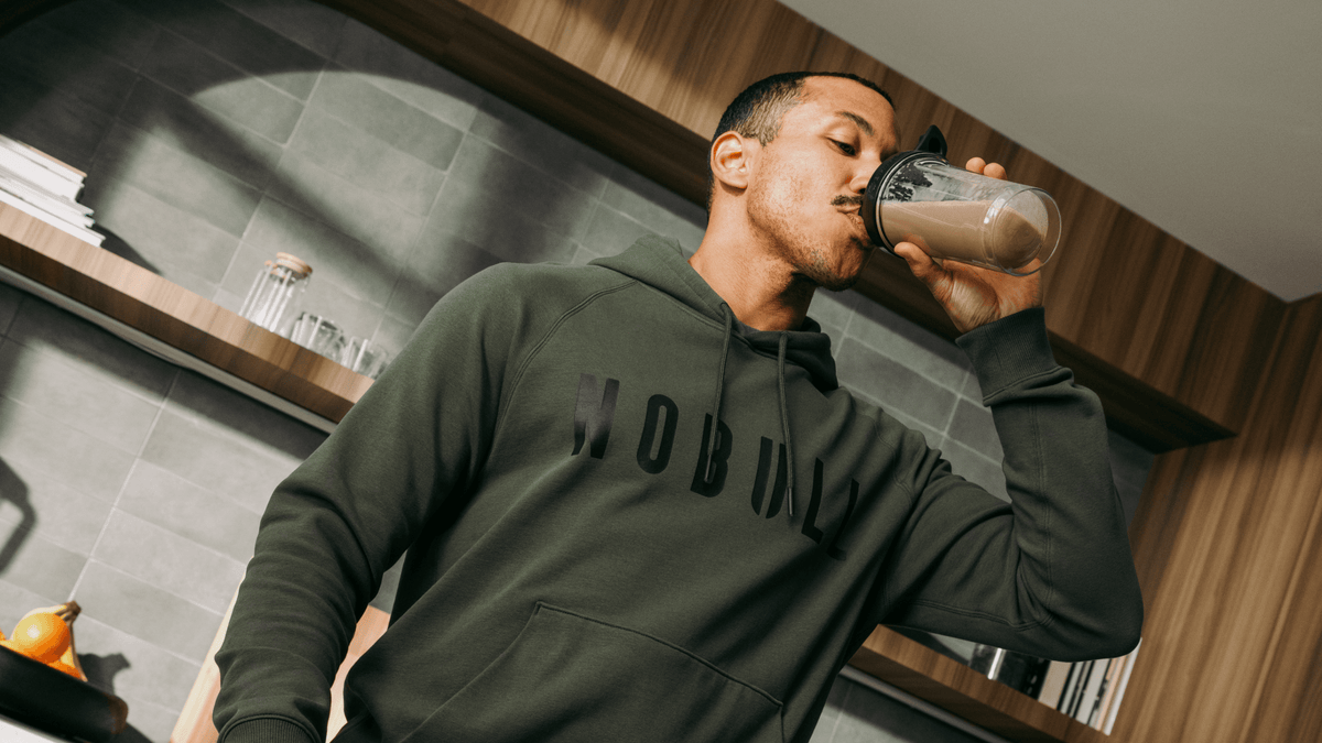 Man drinking chocolate protein from a blender cup in a kitchen wearing NOBULL layers hoodie.